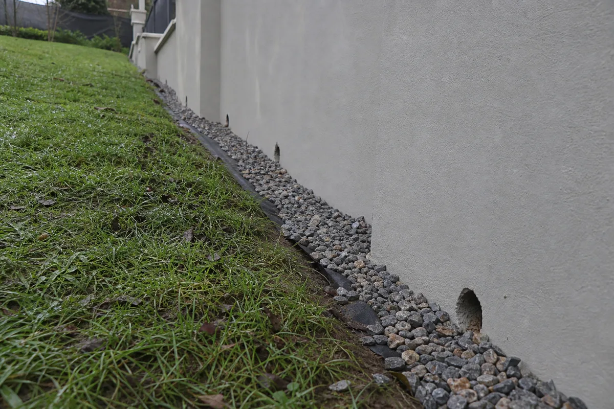 Pied de mur de clôture enduit avec barbacanes d&rsquo;évacuation et lit de graviers sur un jardin en pente